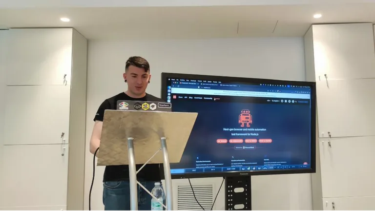 Ludovico Besana speaking as a speaker at the Ministry of Testing Milan meetup, standing behind a lectern with a projector screen in the background.